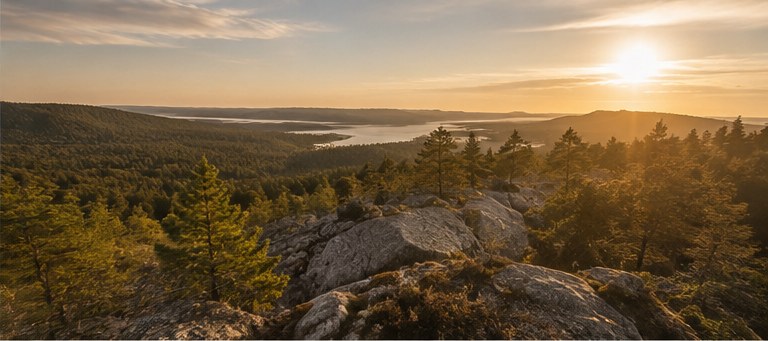 Ljus utsikt över ett vidsträckt sjölandskap i soluppgång, med klippor och skog i förgrunden